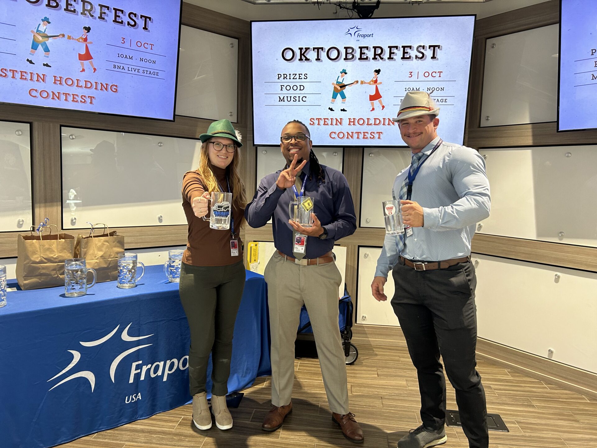 Three people smile holding beer steins at an Oktoberfest stein holding contest at BNA, with event screens and prize table behind them.